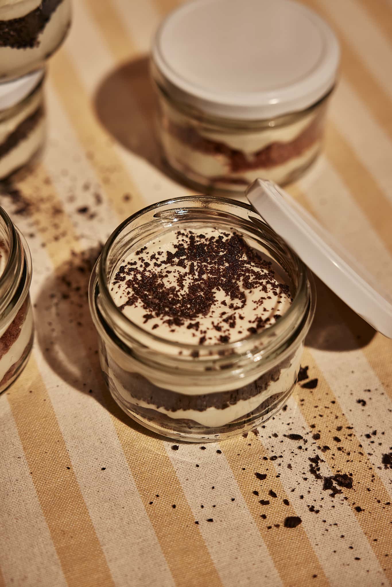 Glass jars filled with layered Oreo tiramisu topped with crushed cookie crumbs on a striped cloth surface.