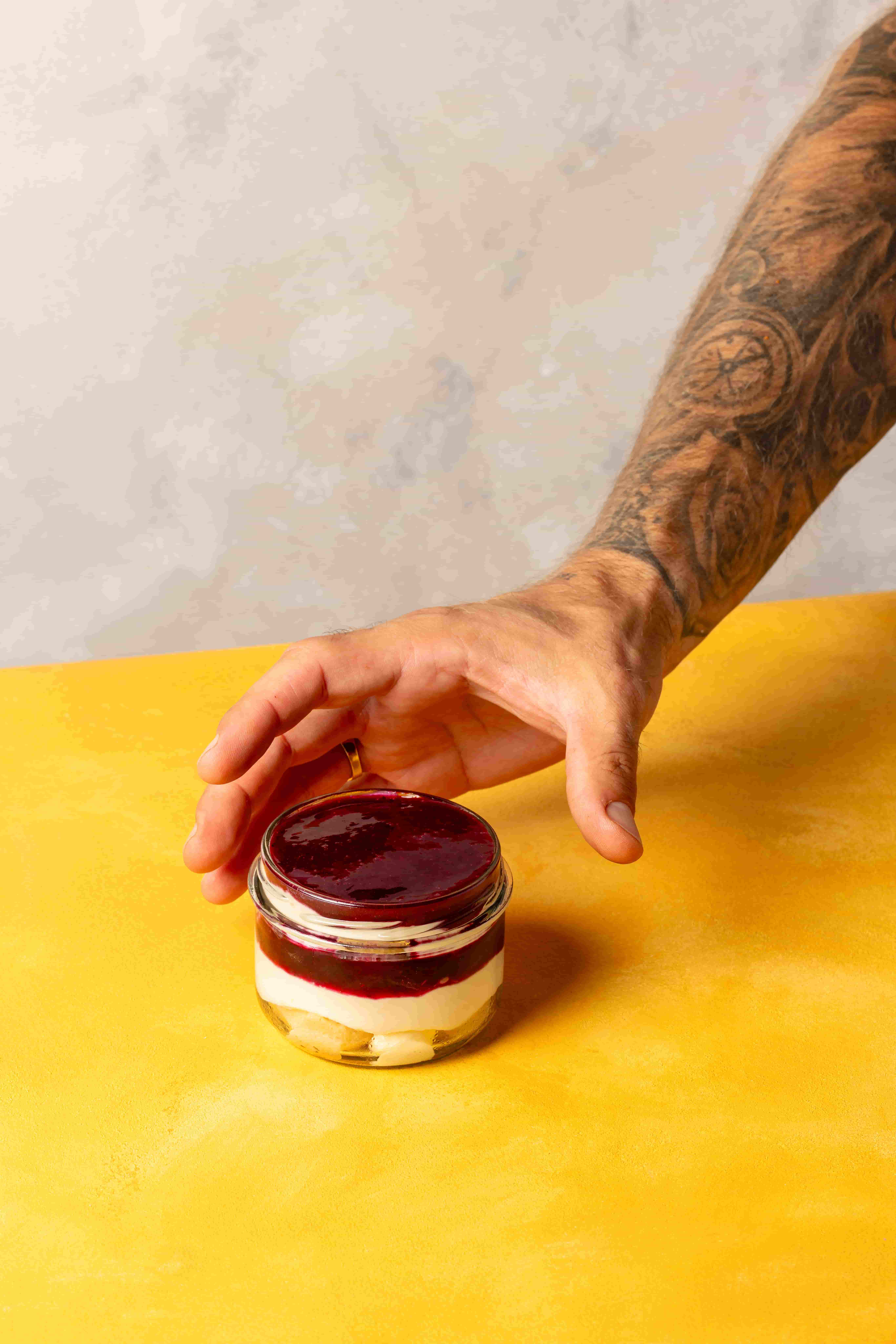 Tattooed arm with a gold ring reaching towards a small glass jar dessert topped with dark red fruit sauce on a yellow surface.
