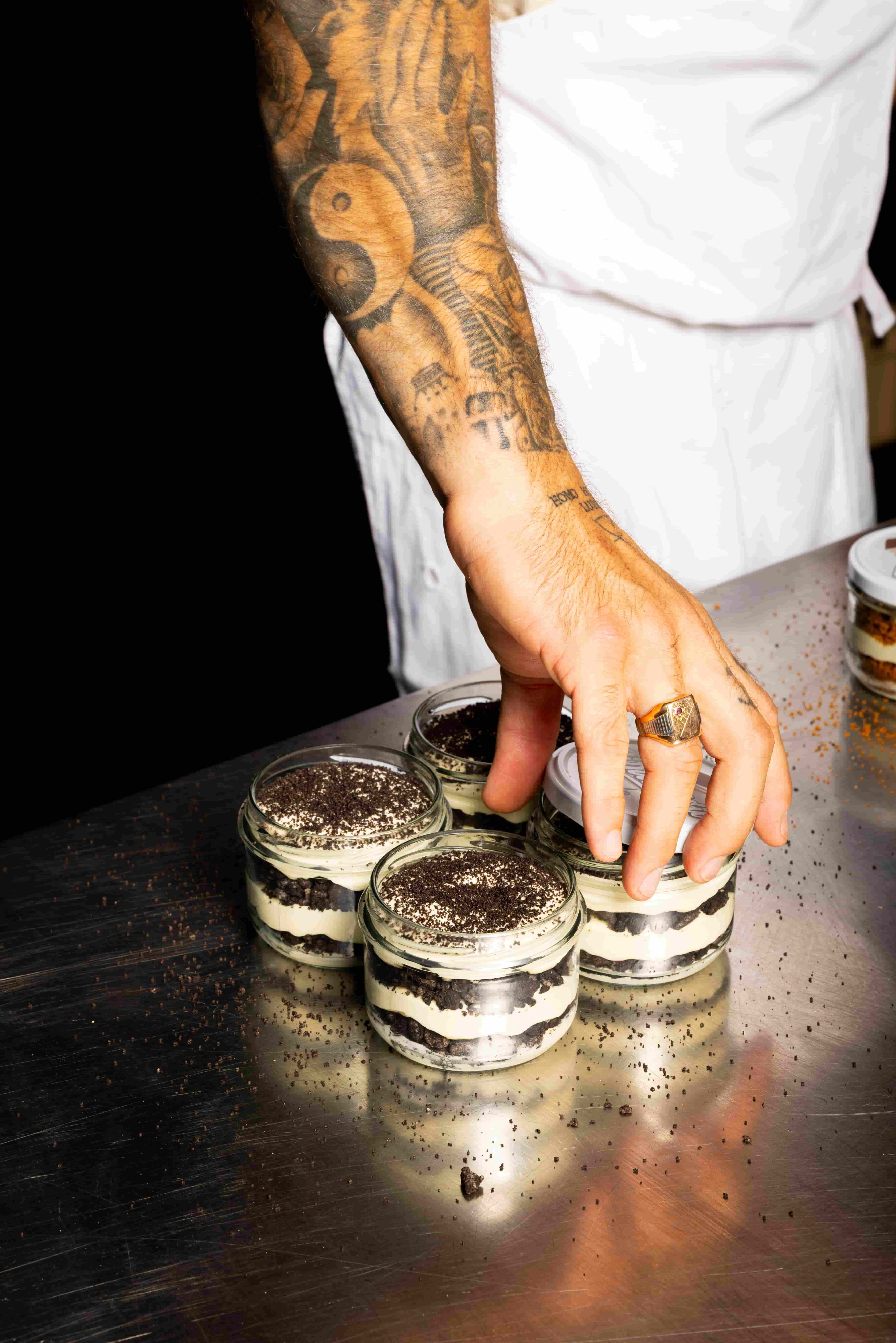 Tattooed hand wearing a ring reaching for one of four glass jars filled with layered Oreo and cream dessert on a metal surface.