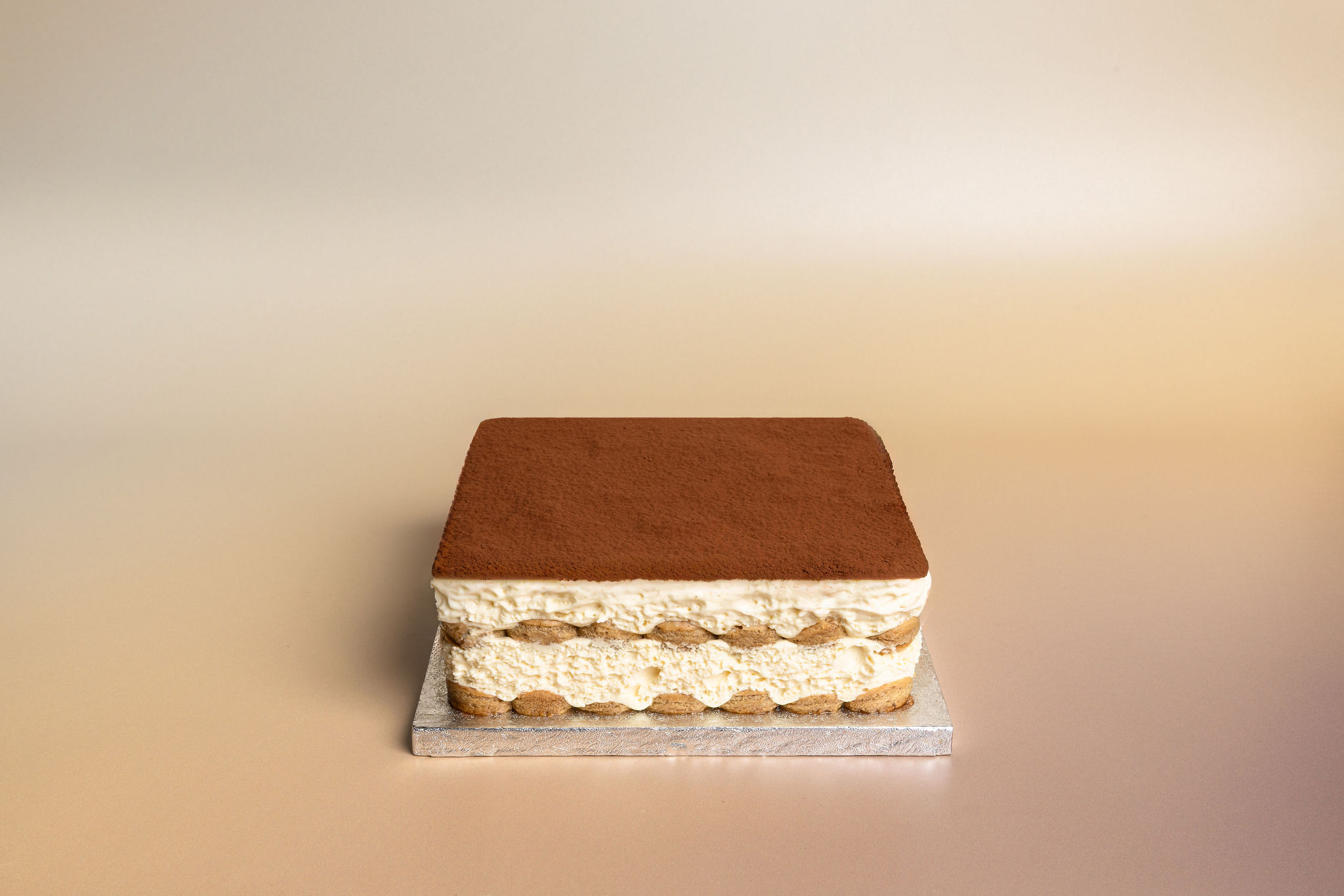 Square tiramisu cake with layers of cream and coffee-soaked biscuits topped with cocoa powder on a silver base.