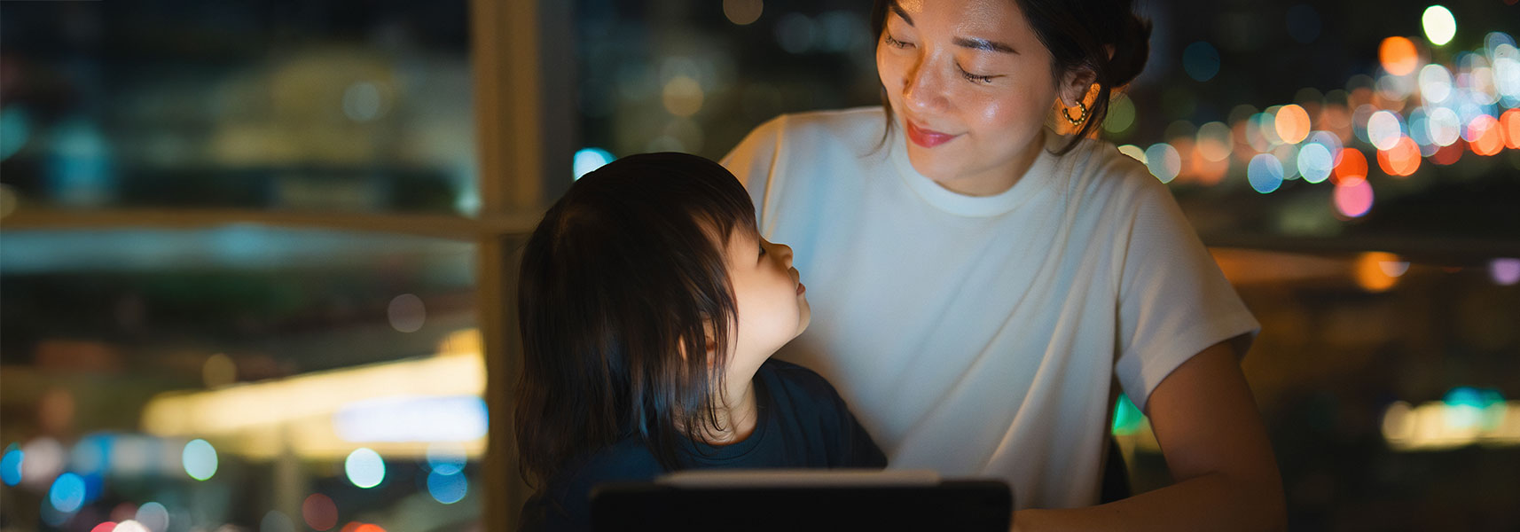 An adult woman sits beside a young child indoors at night. The child looks up at the woman while she looks down at the child with a gentle expression. A tablet or laptop sits in front of them, and blurred city lights glow through a window in the background