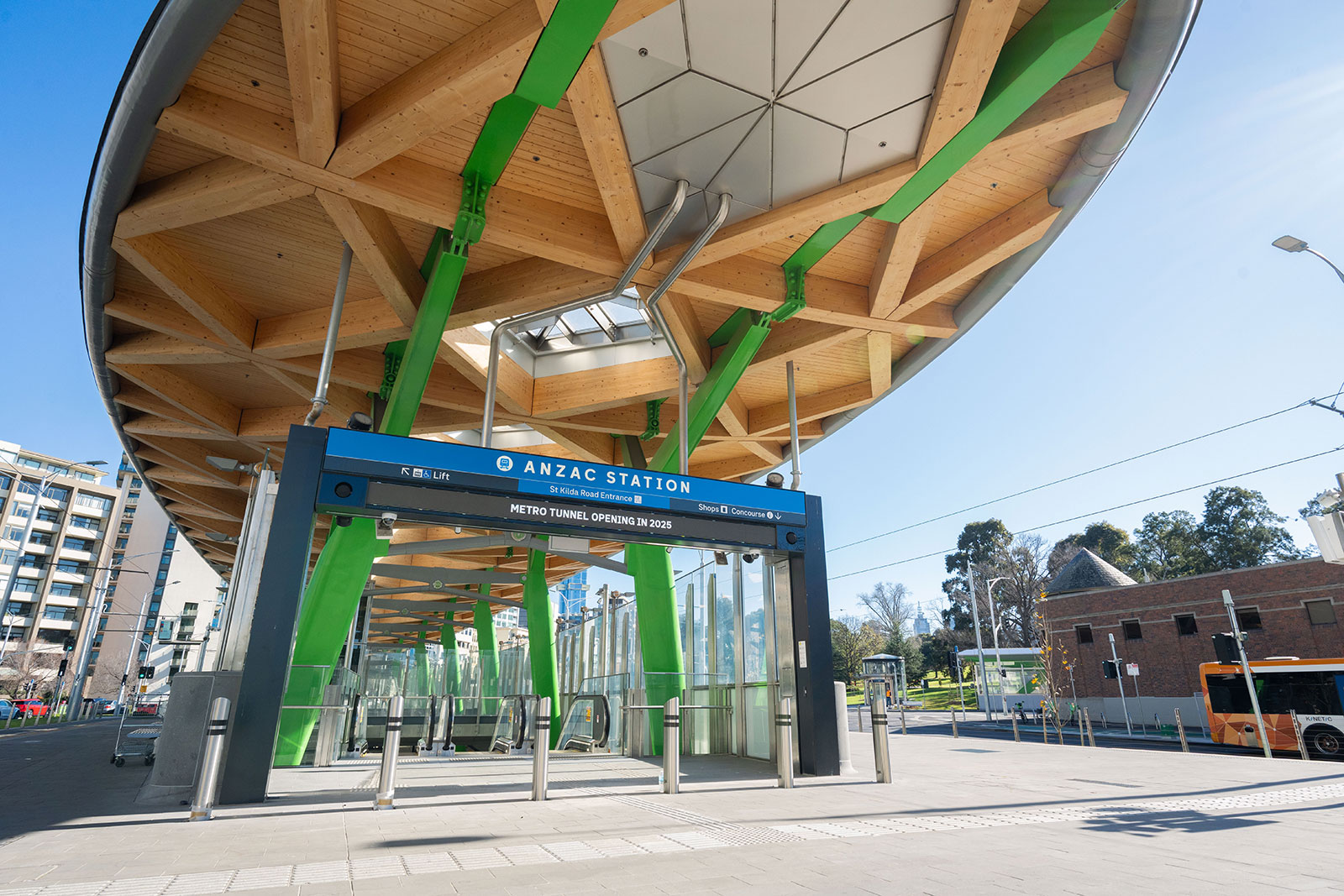 Entrance to ANZAC Station with wooden and green structural canopy and escalators under clear blue sky.