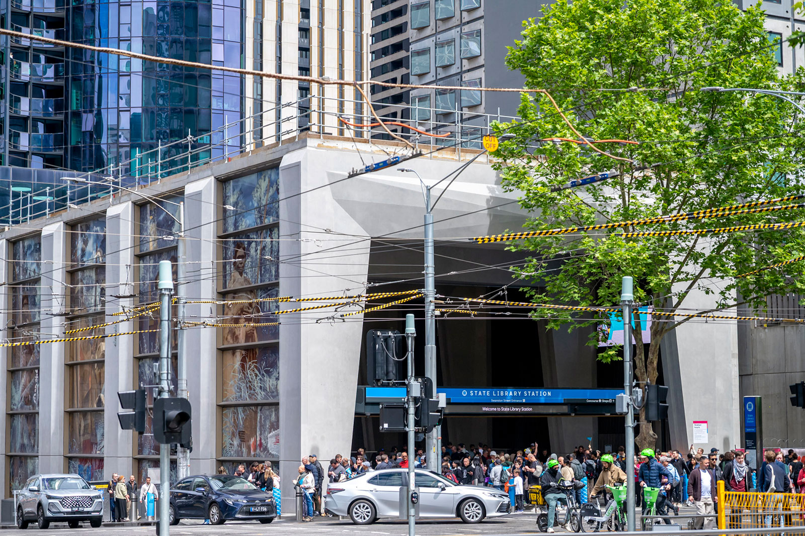 Crowd of people and vehicles in front of the State Library Station entrance in a city street with tall buildings and a large green tree.