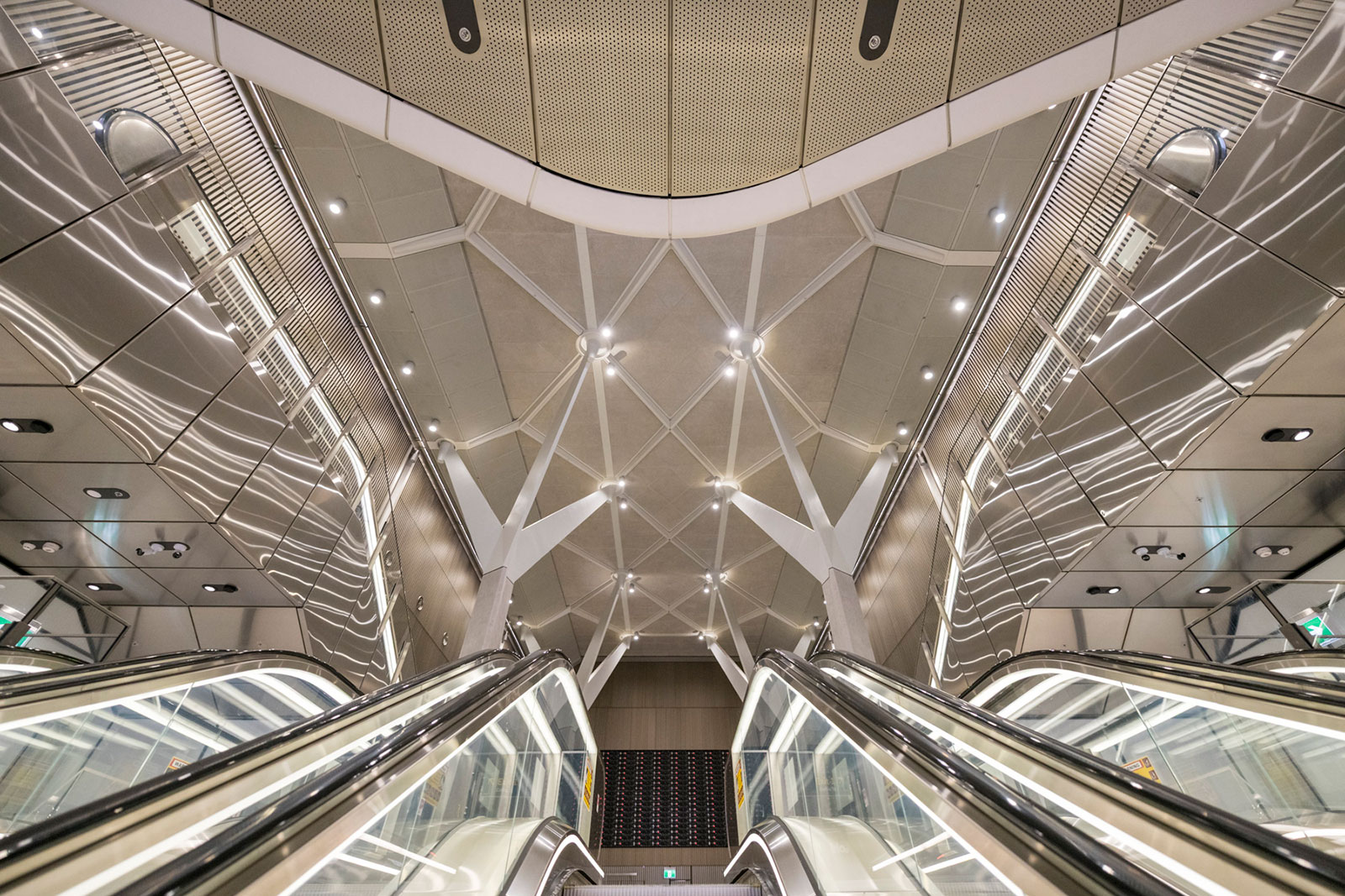 Upward view of a modern escalator flanked by reflective metallic walls and a geometric ceiling with lights.