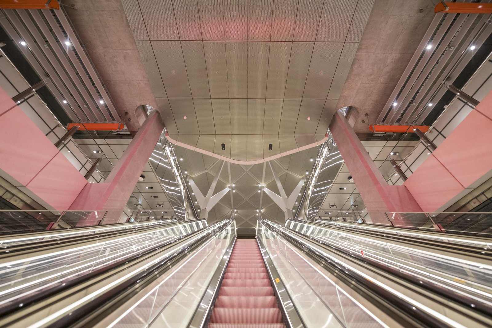 Upward view of red escalators in a modern train station with exposed concrete pillars and geometric ceiling.