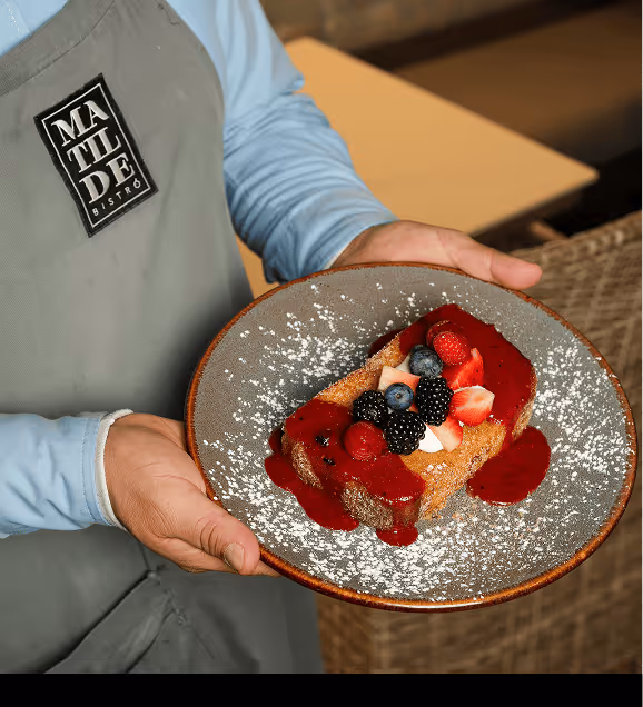 Server wearing a Matilde Bistro apron holding a plate with a dessert topped with mixed berries and red sauce, dusted with powdered sugar.
