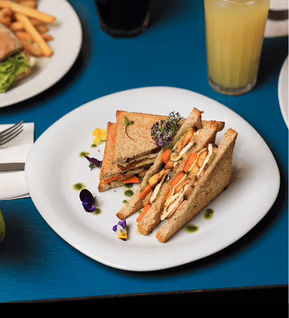 Plate with two triangular sandwiches filled with grilled chicken and carrot slices, garnished with edible flowers and sauce drops, on a blue table.