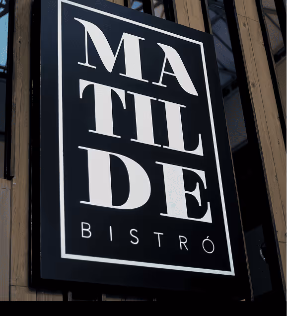 Black sign with white letters spelling 'MATILDE BISTRÓ' mounted on a wooden slat wall.