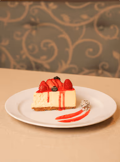 Slice of cheesecake topped with strawberries, raspberries, blueberries, and a red sauce drizzle, served on a white plate with a decorative sauce swirl and a small garnish.