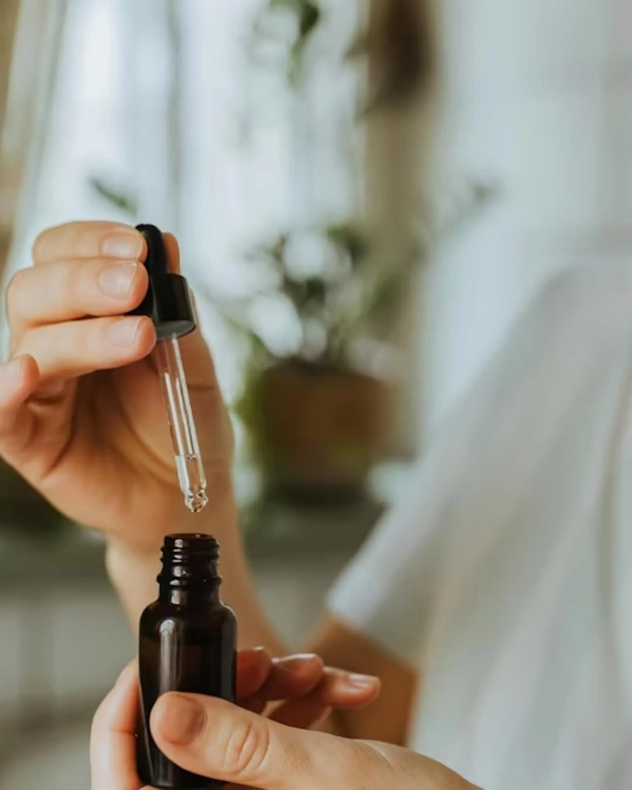 Person holding a dropper above a small dark glass bottle with a blurred indoor background.
