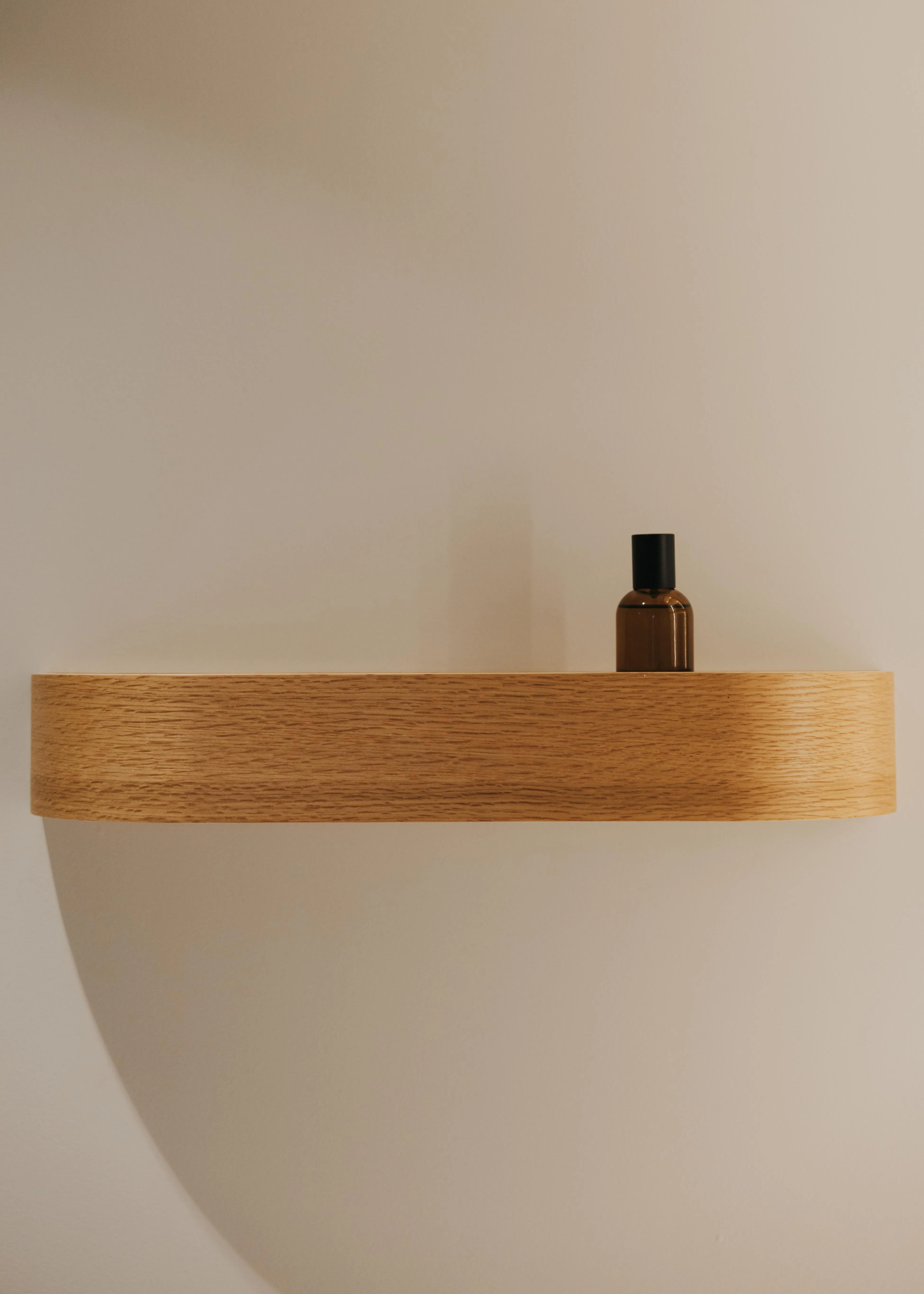 Minimalist wooden floating shelf mounted on a light-colored wall with a single small amber bottle with a black cap on it.