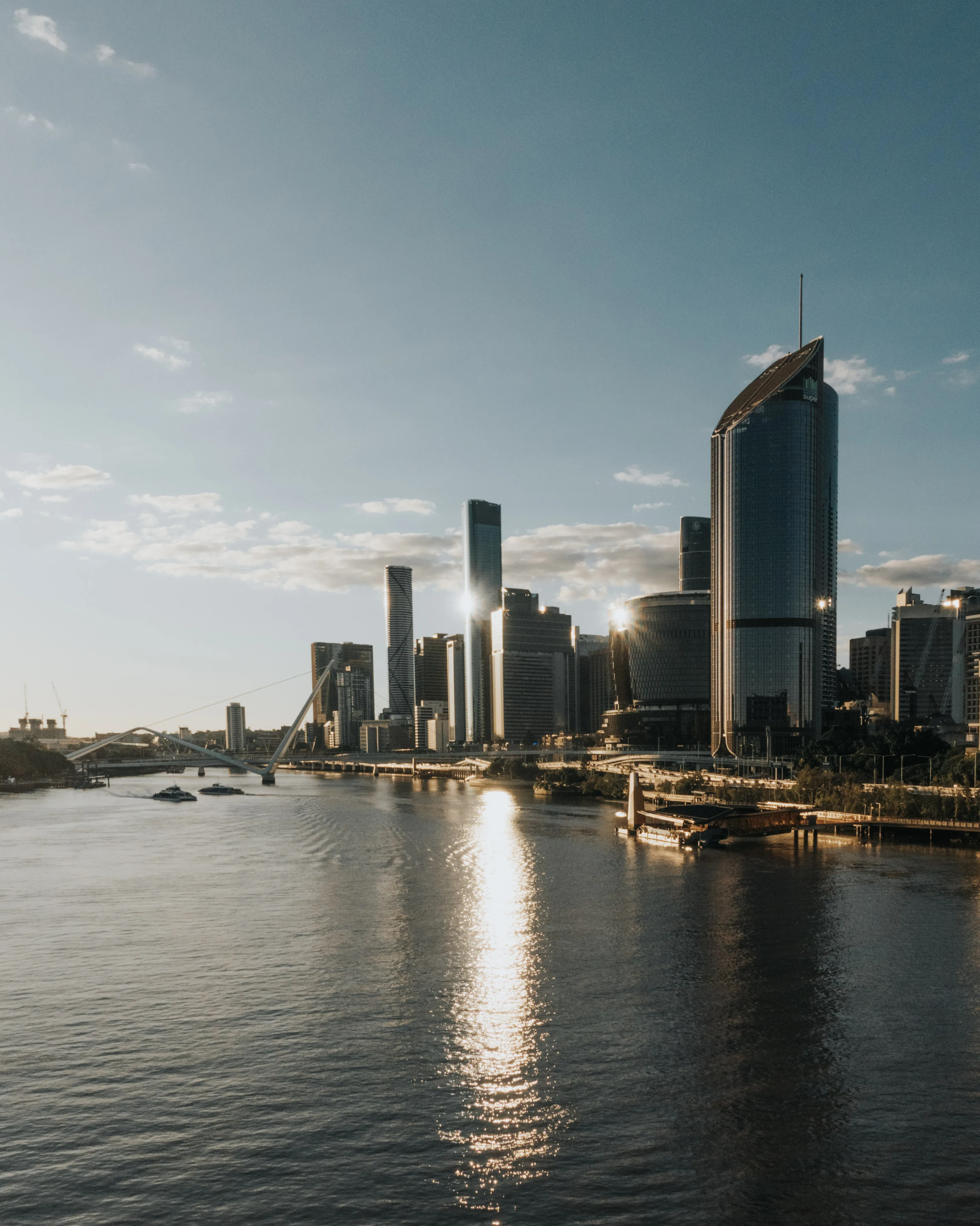 City skyline with modern skyscrapers reflecting sunlight by a wide river under a partly cloudy sky.