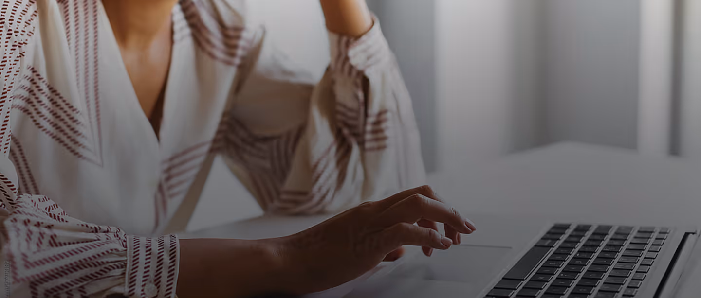 Person in patterned shirt using a laptop on a white desk in a softly lit room.