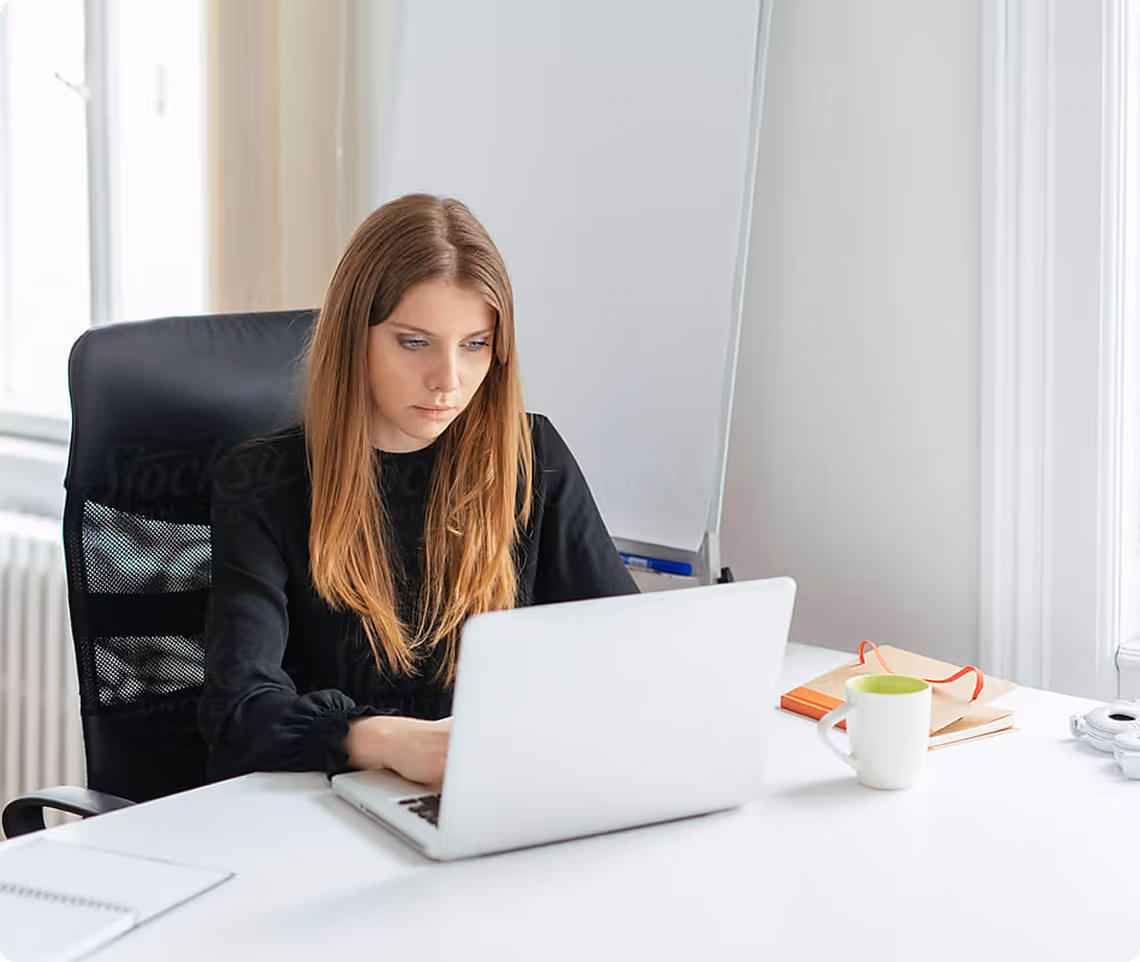 Young woman with long hair working on a laptop at a white desk in a bright office.