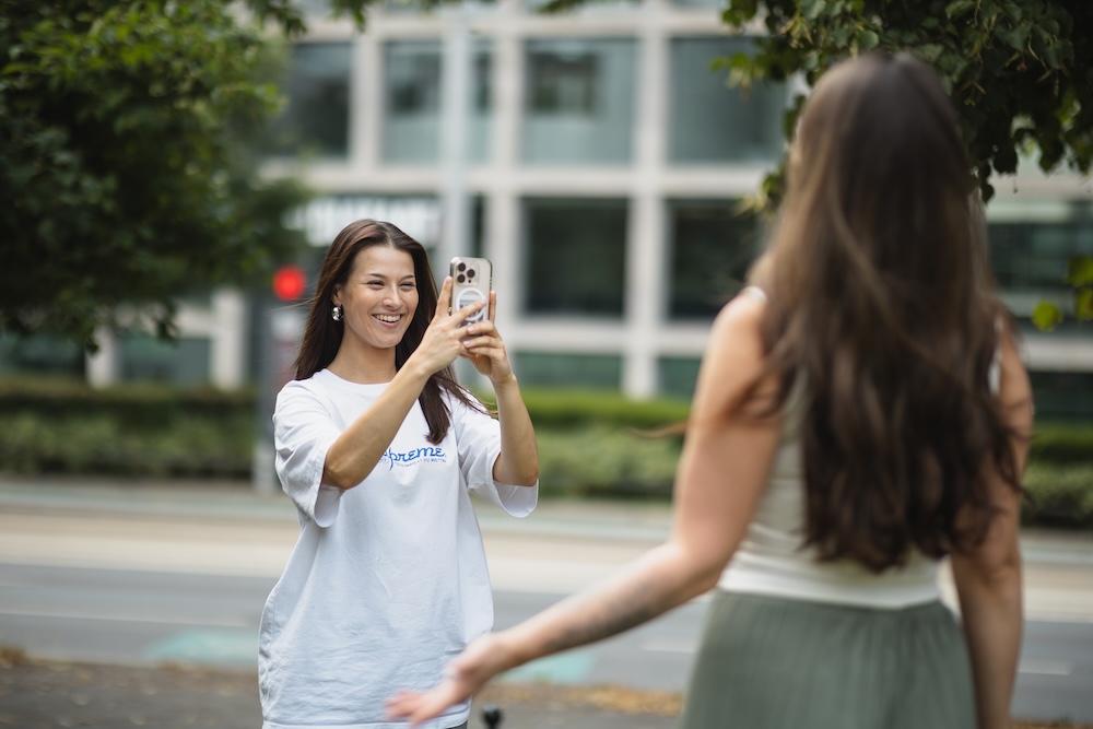 Frau in weißem T-Shirt macht ein Foto mit ihrem Smartphone von einer anderen Frau draußen.
