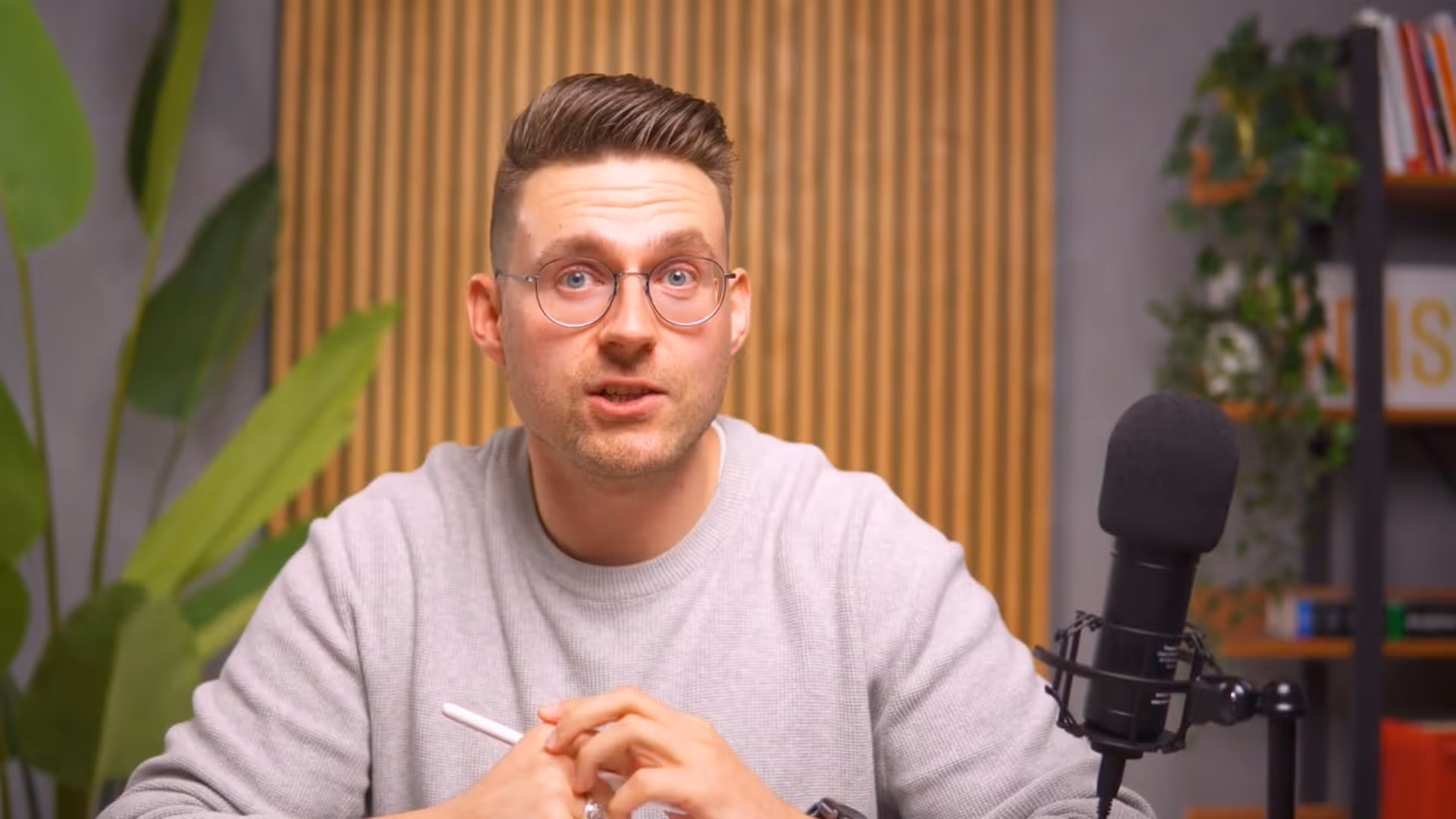 Mann mit Brille spricht vor Mikrofon vor Holzpaneelen und Pflanzen im Hintergrund.