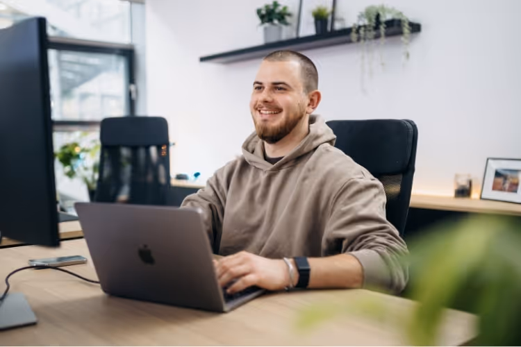 Junger Mann in Hoodie lächelt, sitzt am Schreibtisch mit Laptop und Monitor in modernem Büro.