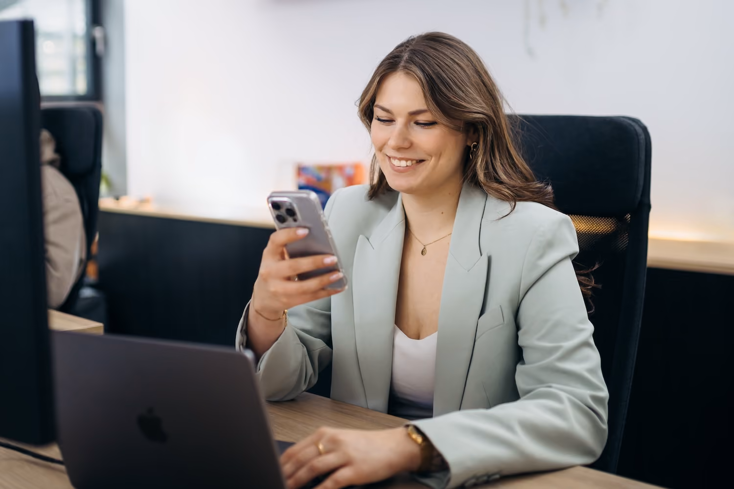 Frau in Büro mit Laptop lächelt und schaut auf ihr Handy.