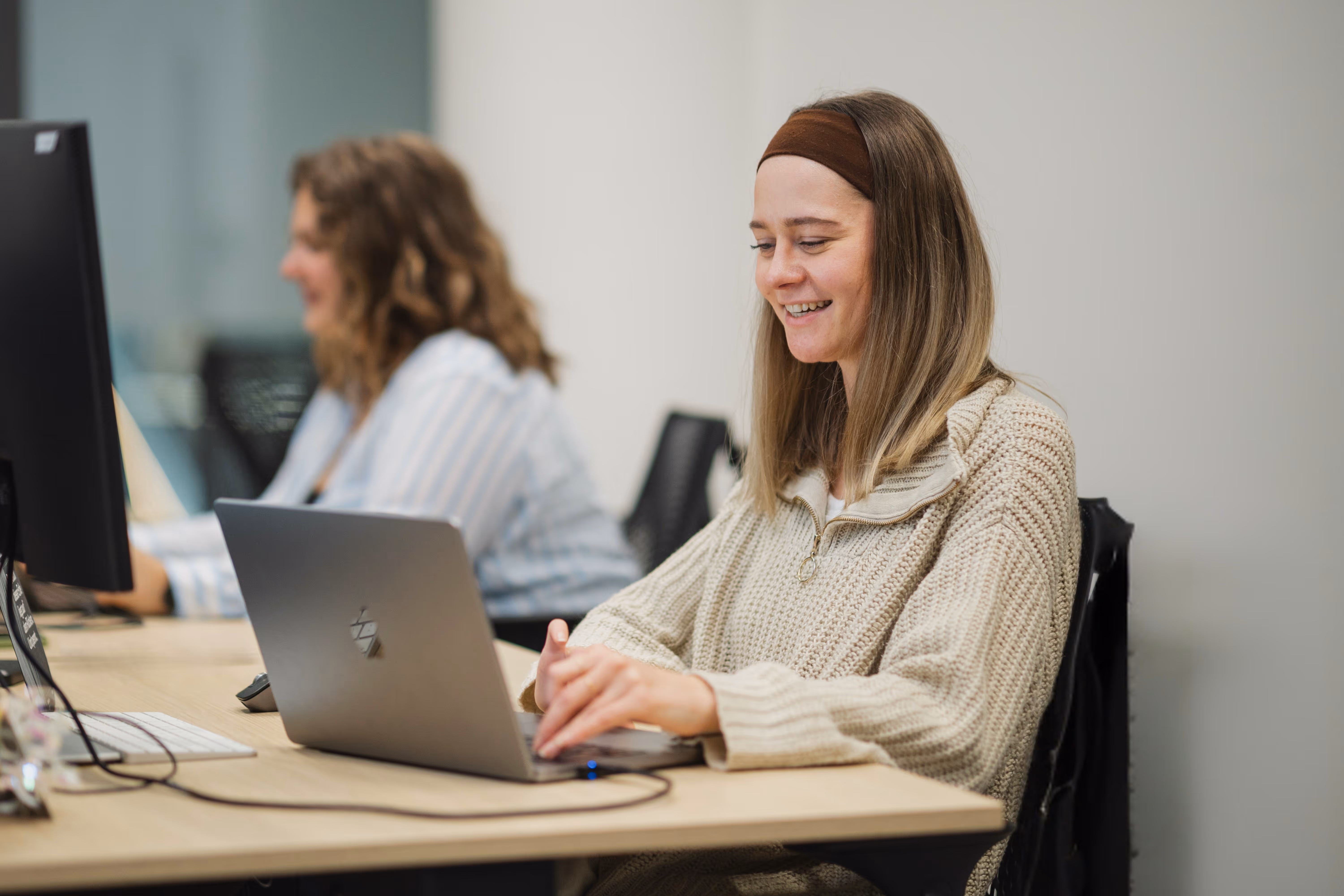 Zwei Frauen arbeiten in einem Büro an Computern, eine lächelt auf ihren Laptop.