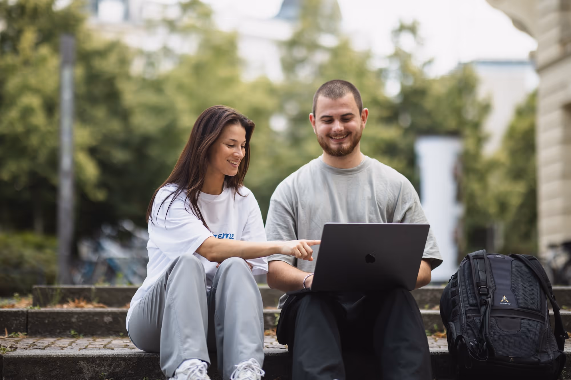 Zwei junge Erwachsene sitzen im Freien auf Stufen, eine Frau zeigt auf den Bildschirm eines Laptops, beide lächeln.