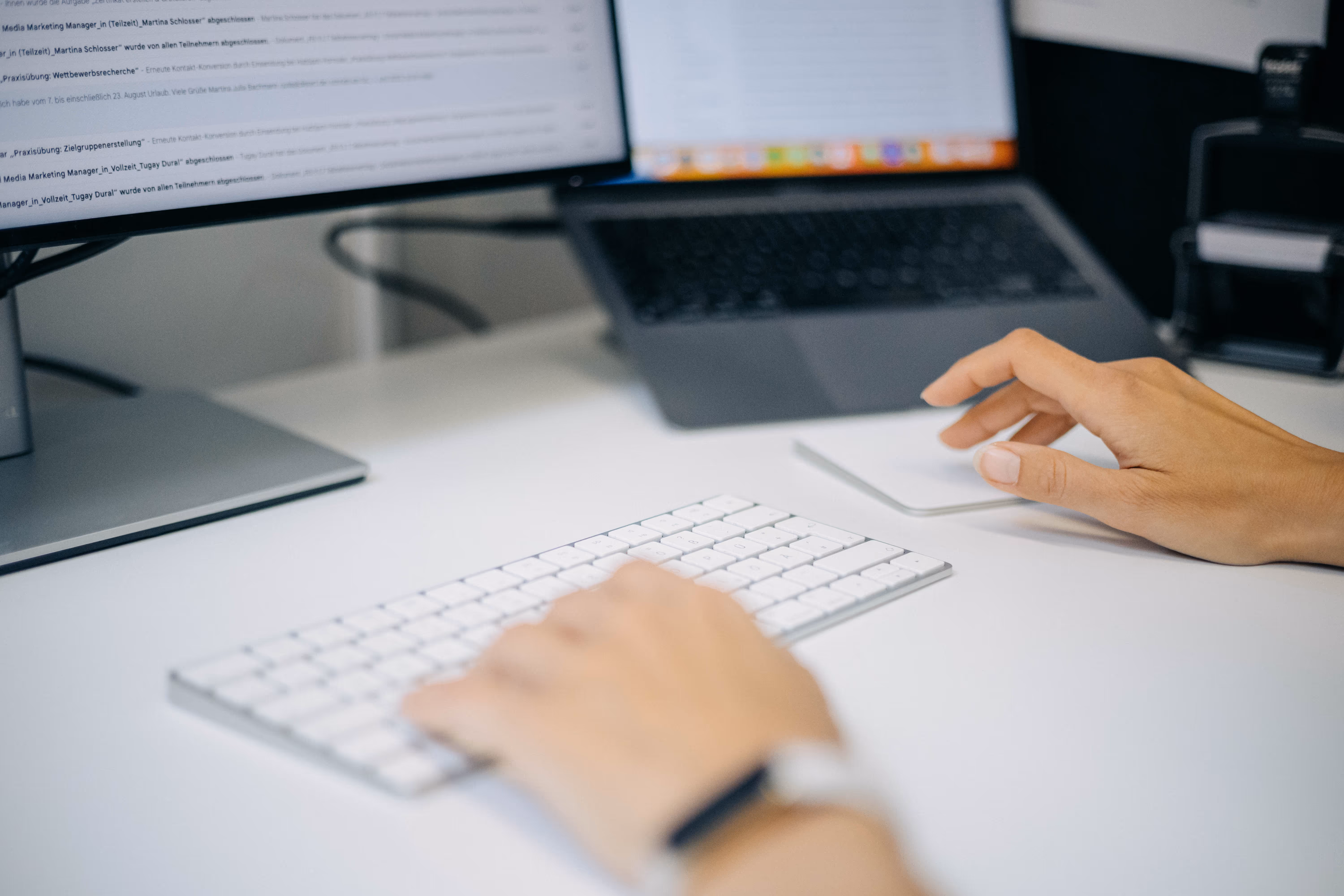 Person arbeitet mit Laptop, Monitor und weißer Tastatur an weißem Schreibtisch.