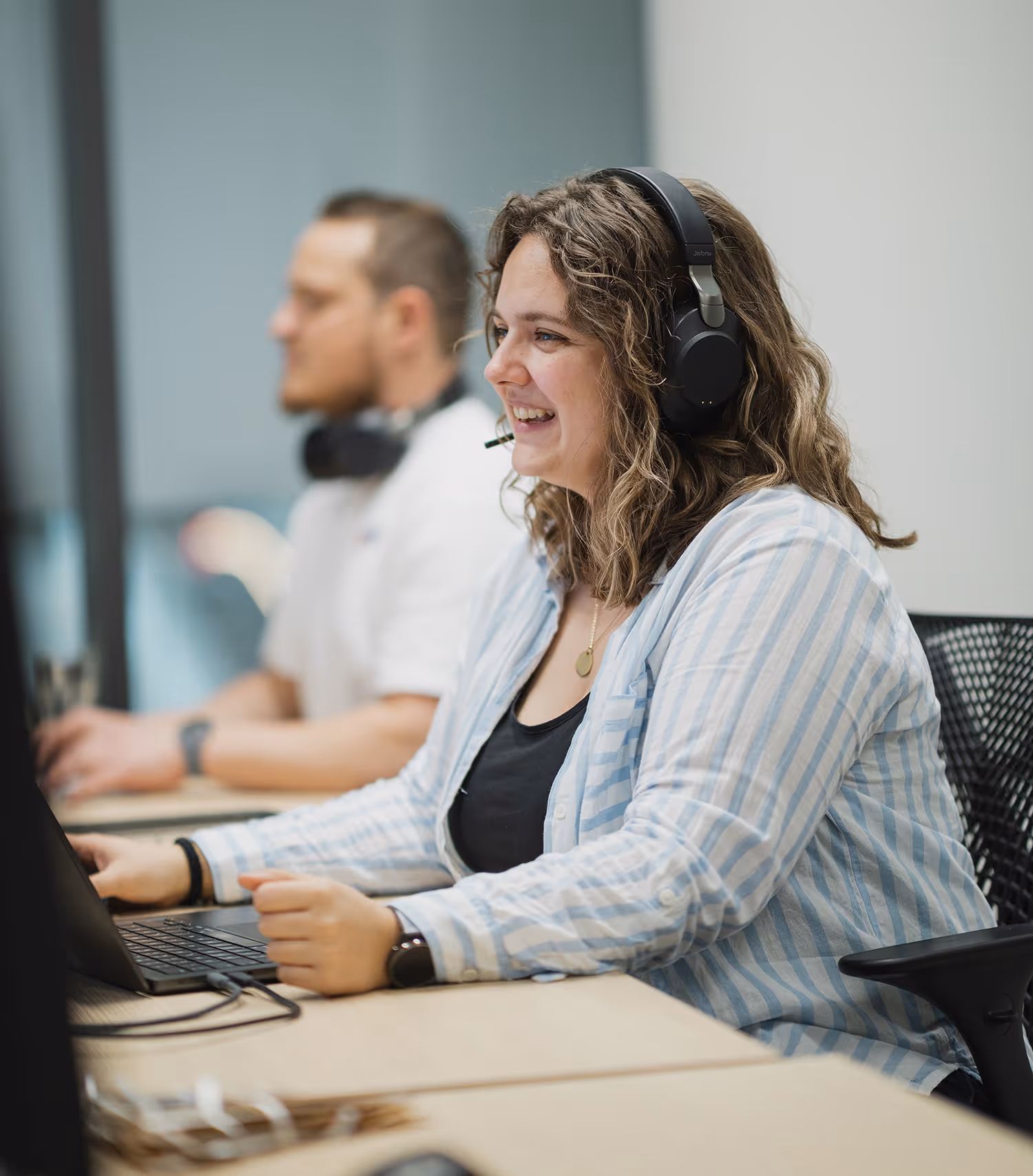 Lächelnde Frau mit lockigem Haar und Headset arbeitet an einem Laptop in einem Büro.