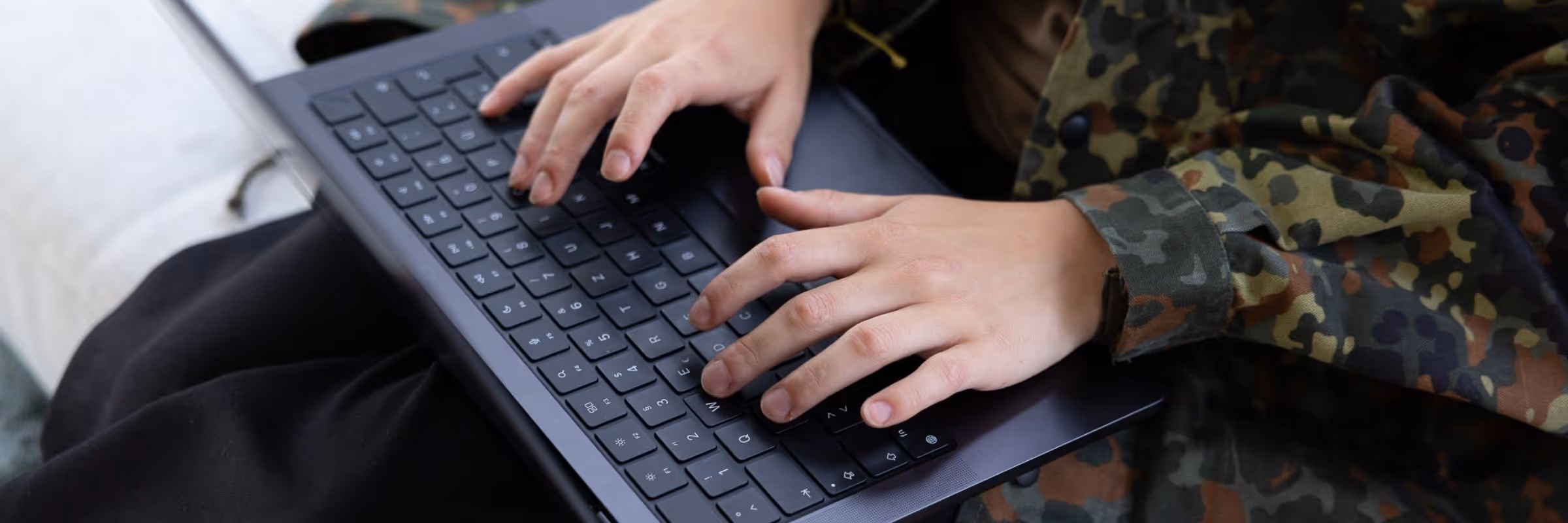 Hände tippen auf schwarze Laptop-Tastatur, Person trägt Camouflage-Jacke.