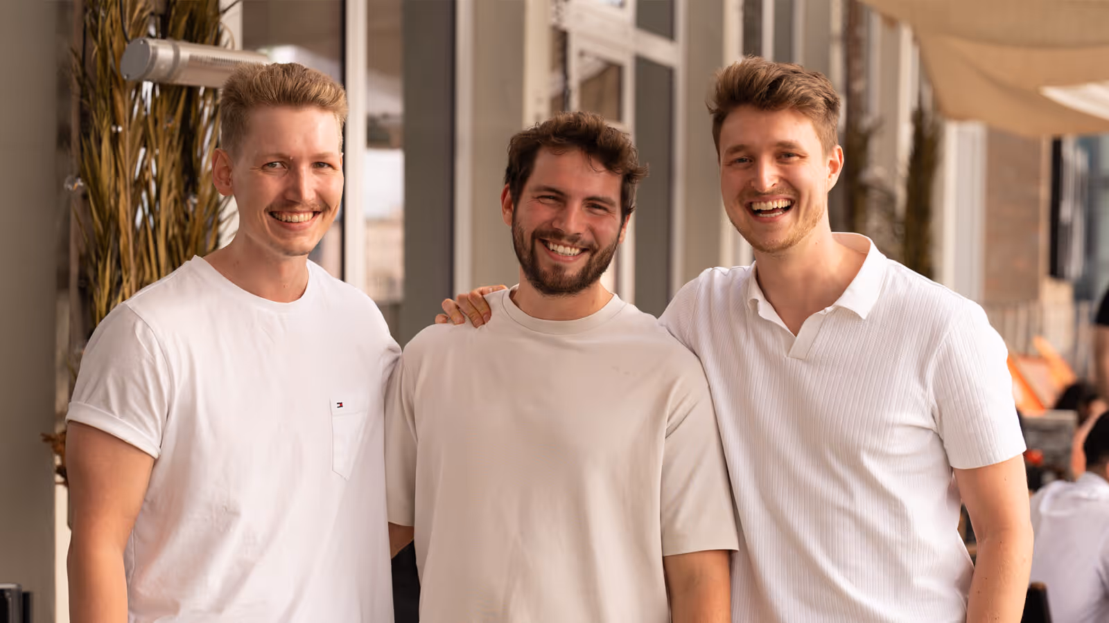 Drei lächelnde junge Männer in hellen T-Shirts stehen nah beieinander und posieren in einem modernen Innenraum.