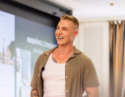 Junger Mann mit kurzen blonden Haaren und Tattoo spricht lächelnd vor einem Bildschirm in einem modernen Raum.