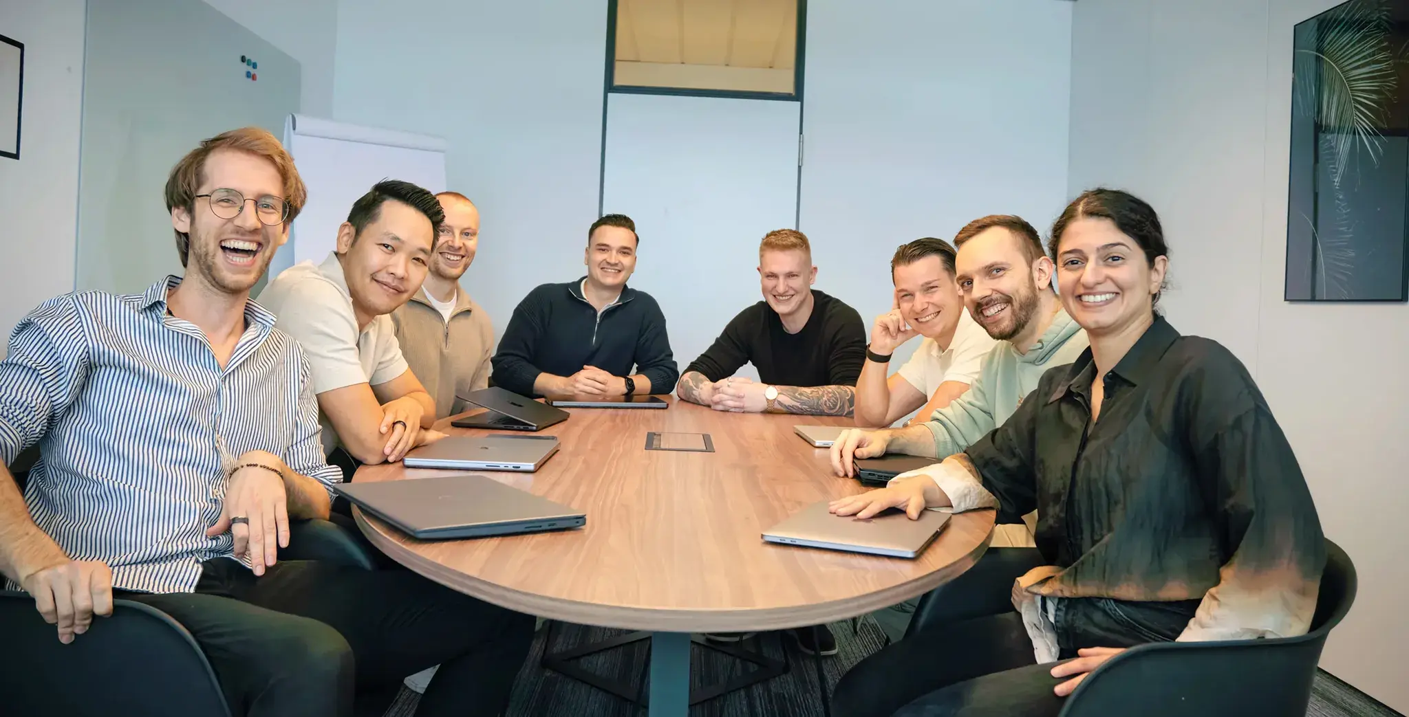 Eine diverse Gruppe von acht Menschen, die um einen Konferenztisch sitzen und in einem Büro lächeln.