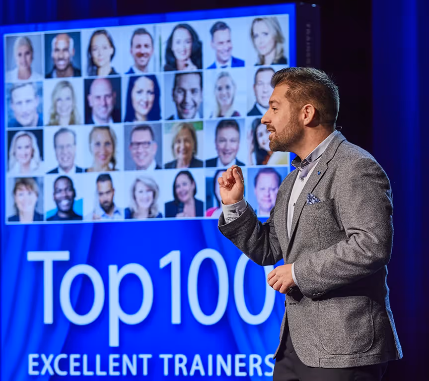 Ein Mann im grauen Sakko spricht auf einer Bühne vor einer großen Anzeige mit den Gesichtern vieler Menschen und dem Text 'Top 100 Excellent Trainers'.