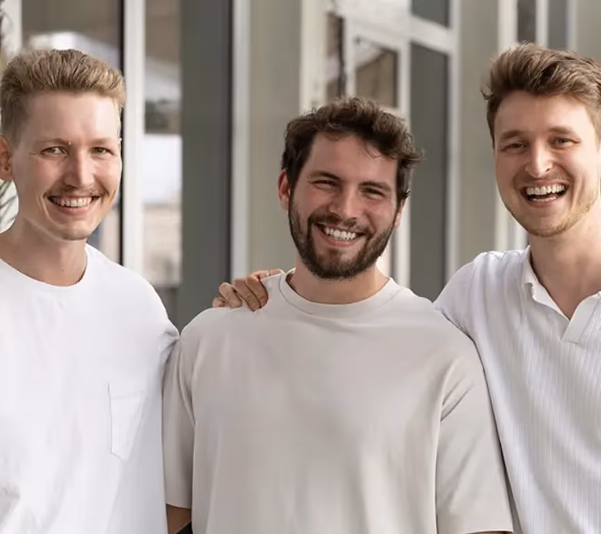 Drei Männer in weißem T-Shirt lächeln und posieren gemeinsam vor einem Fenster.