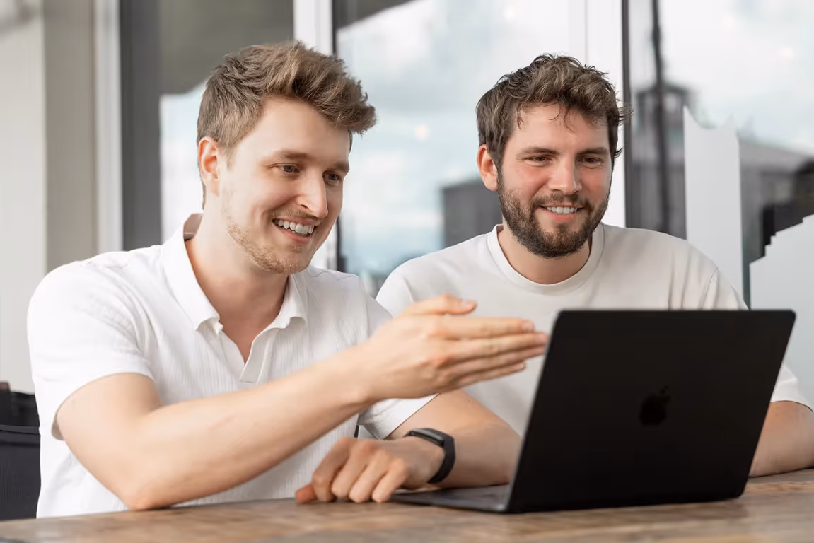 Zwei Männer lächeln und schauen zusammen auf einen schwarzen Laptop in einem hellen Büro.