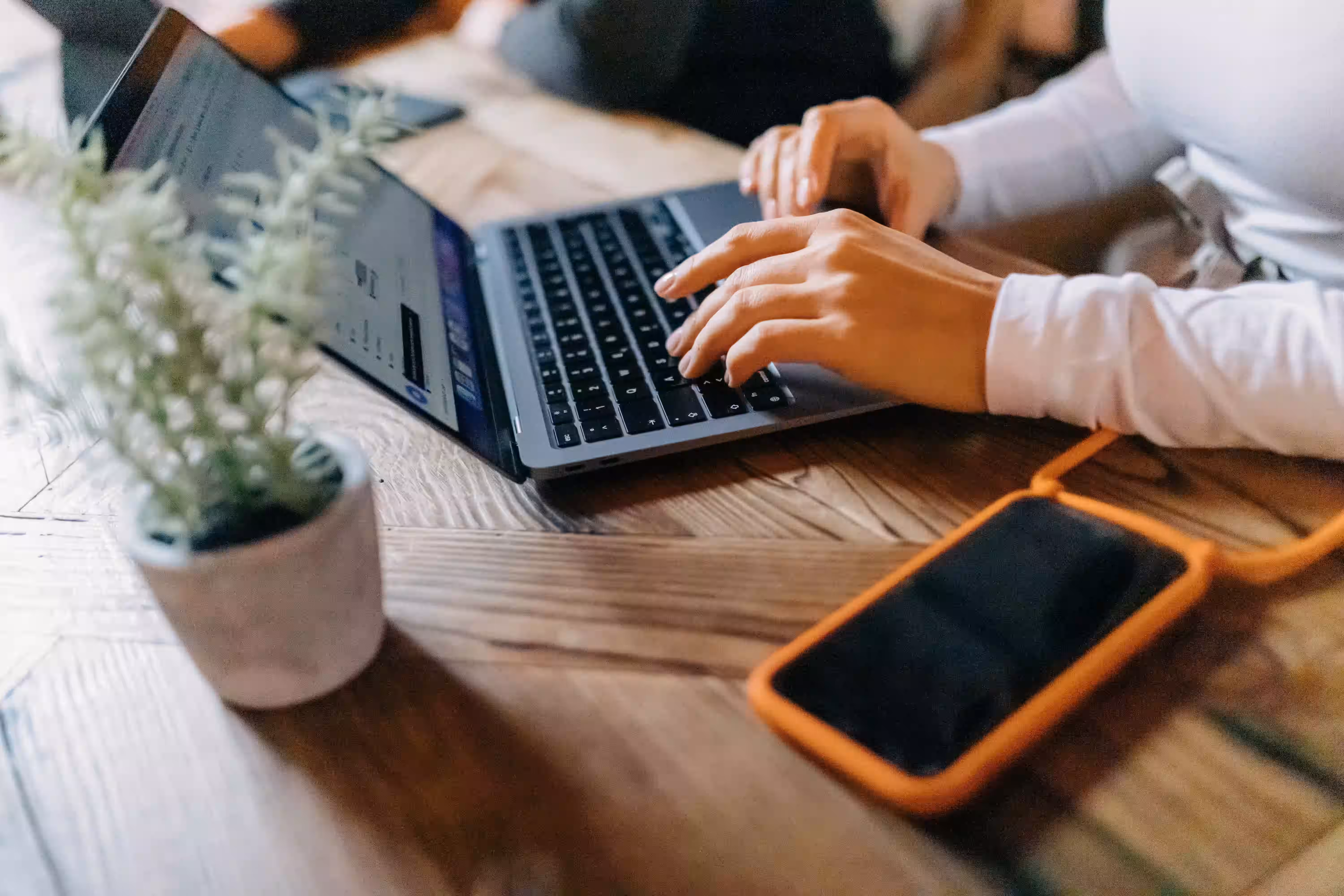 Person tippt auf einem Laptop, daneben liegt ein Smartphone mit orangefarbenem Schutzcase auf einem Holztisch.