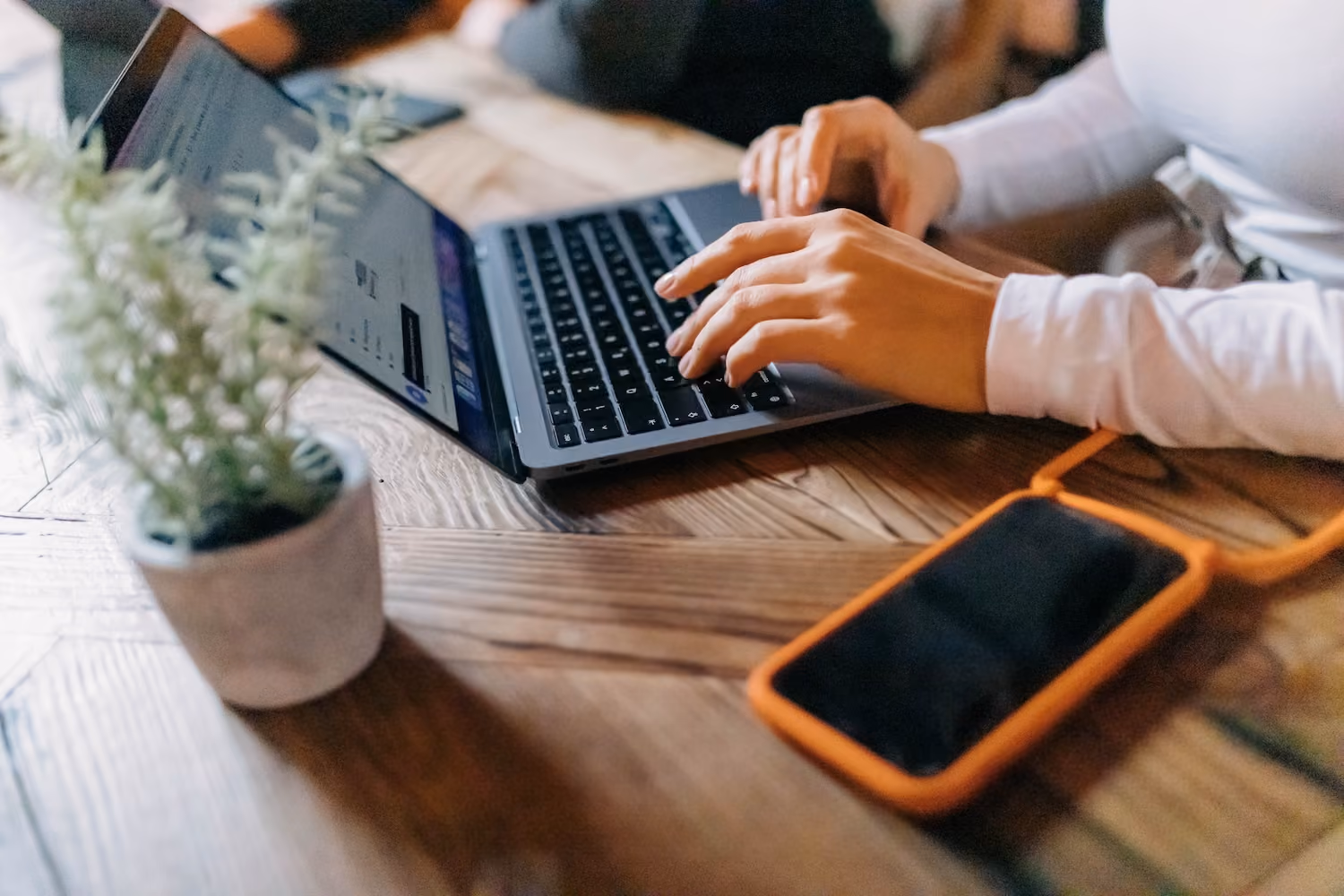 Person tippt auf einem Laptop auf einem Holztisch mit Smartphone und Pflanze daneben.