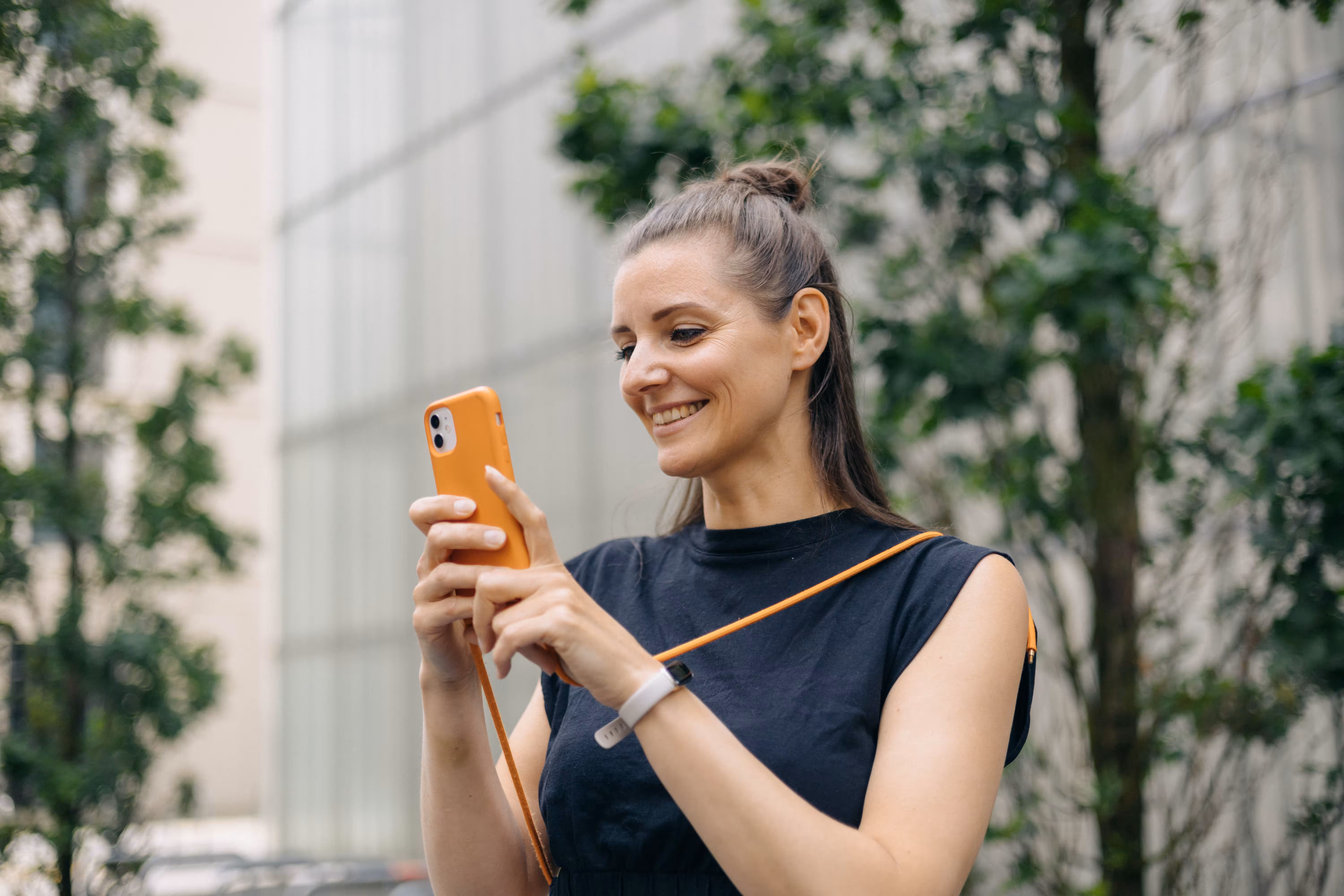 Lächelnde Frau mit hochgebundenem Haar benutzt im Freien ein Smartphone mit orangem Schutzcase.