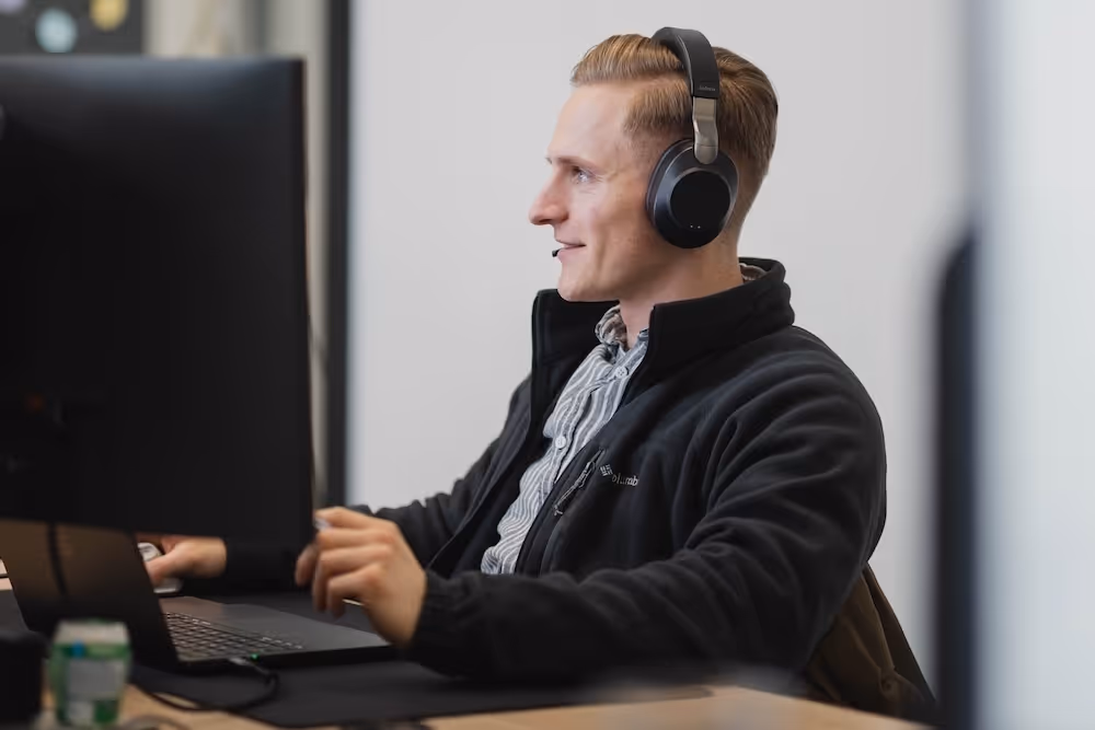 Junger Mann mit Kopfhörern arbeitet am Computer an einem Schreibtisch.
