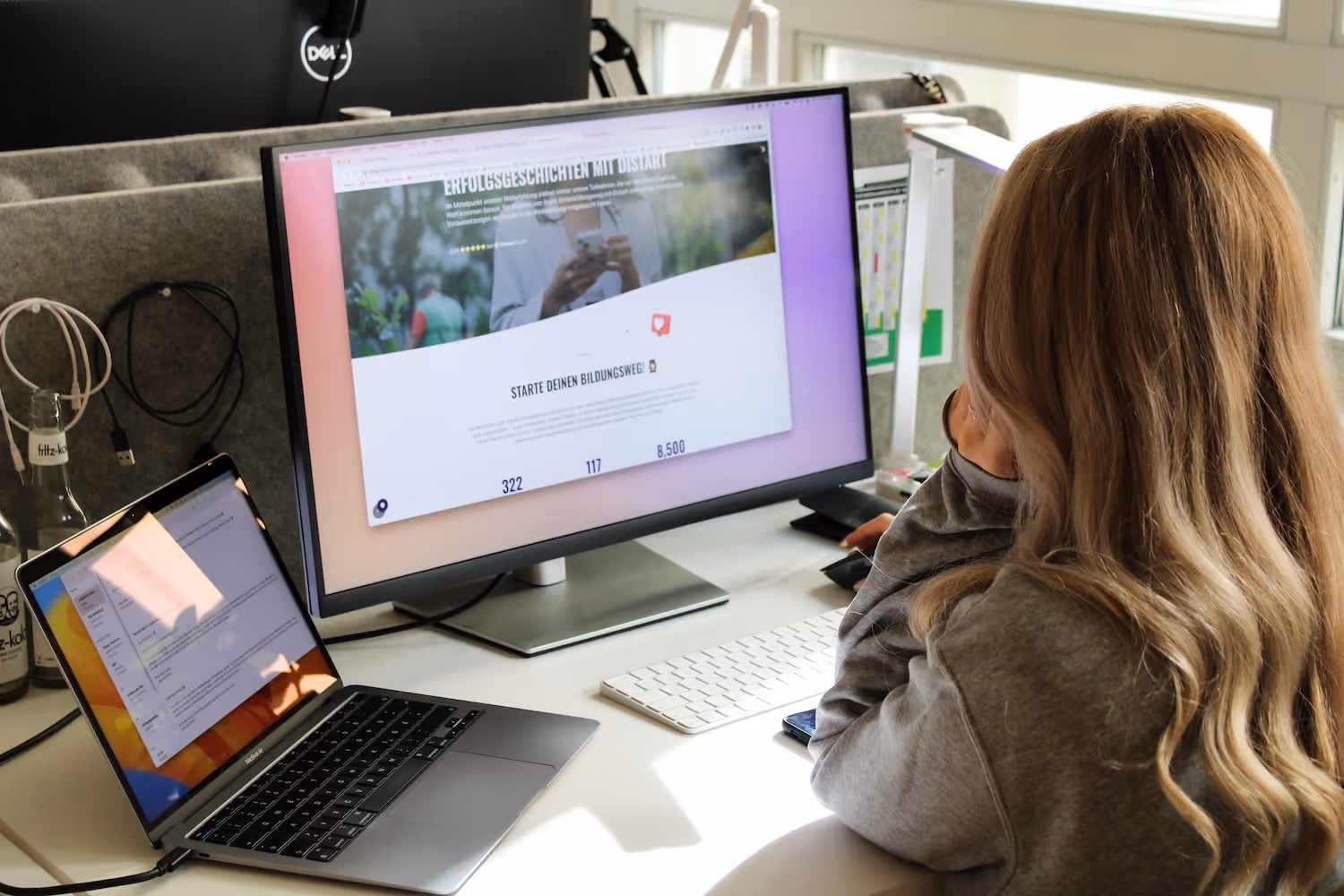 Frau mit langen blonden Haaren arbeitet an einem Schreibtisch mit Laptop und großem Monitor.