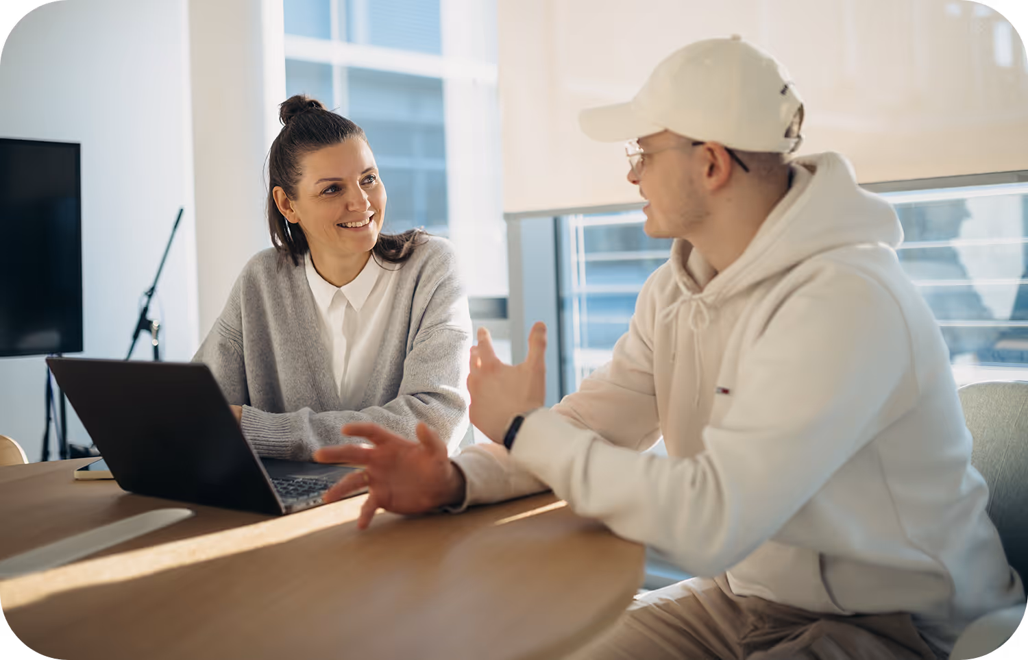 Frau im grauen Cardigan und Mann in weißem Hoodie unterhalten sich an einem Tisch mit Laptop.