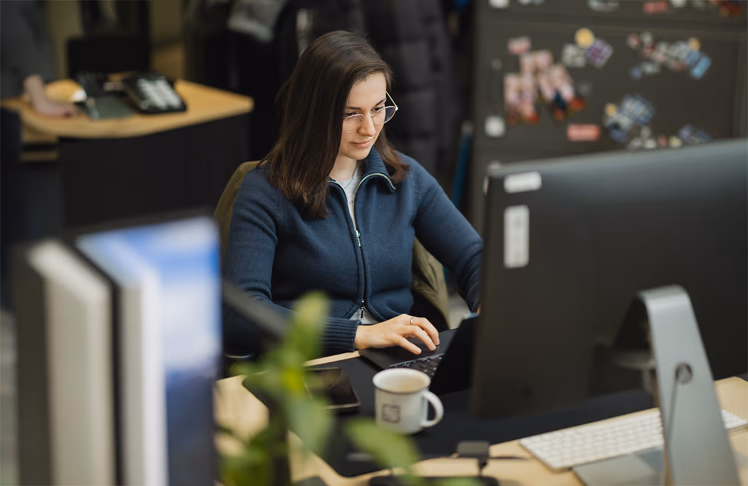 Frau mit Brille arbeitet an Laptop in Büro mit Monitor, Kaffeetasse und Smartphone auf dem Tisch.