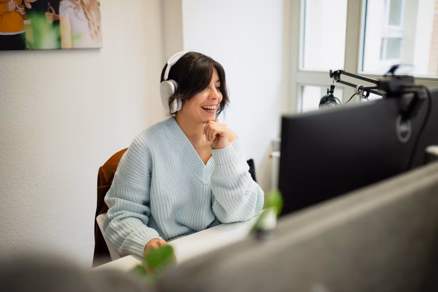 Frau mit Kopfhörern lächelt bei der Arbeit am Computer in einem hellen Büro.