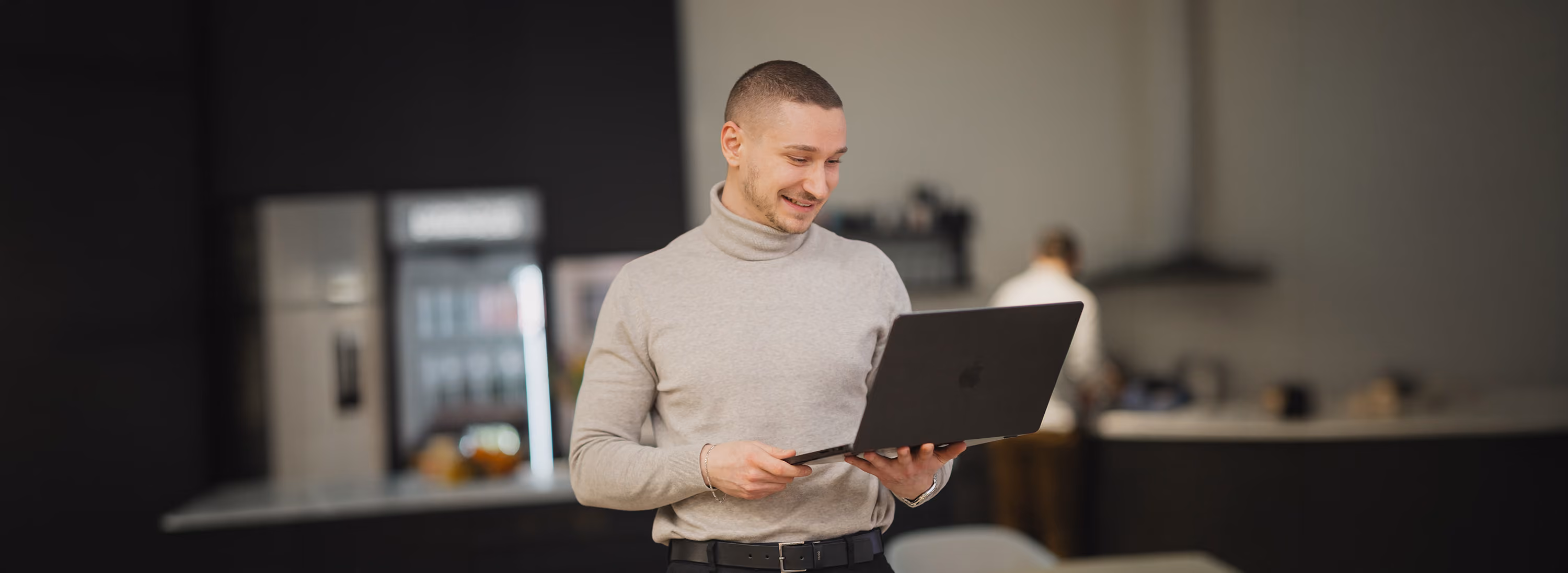 Junger Mann in Rollkragenpullover lächelt und hält Laptop in moderner Küche.