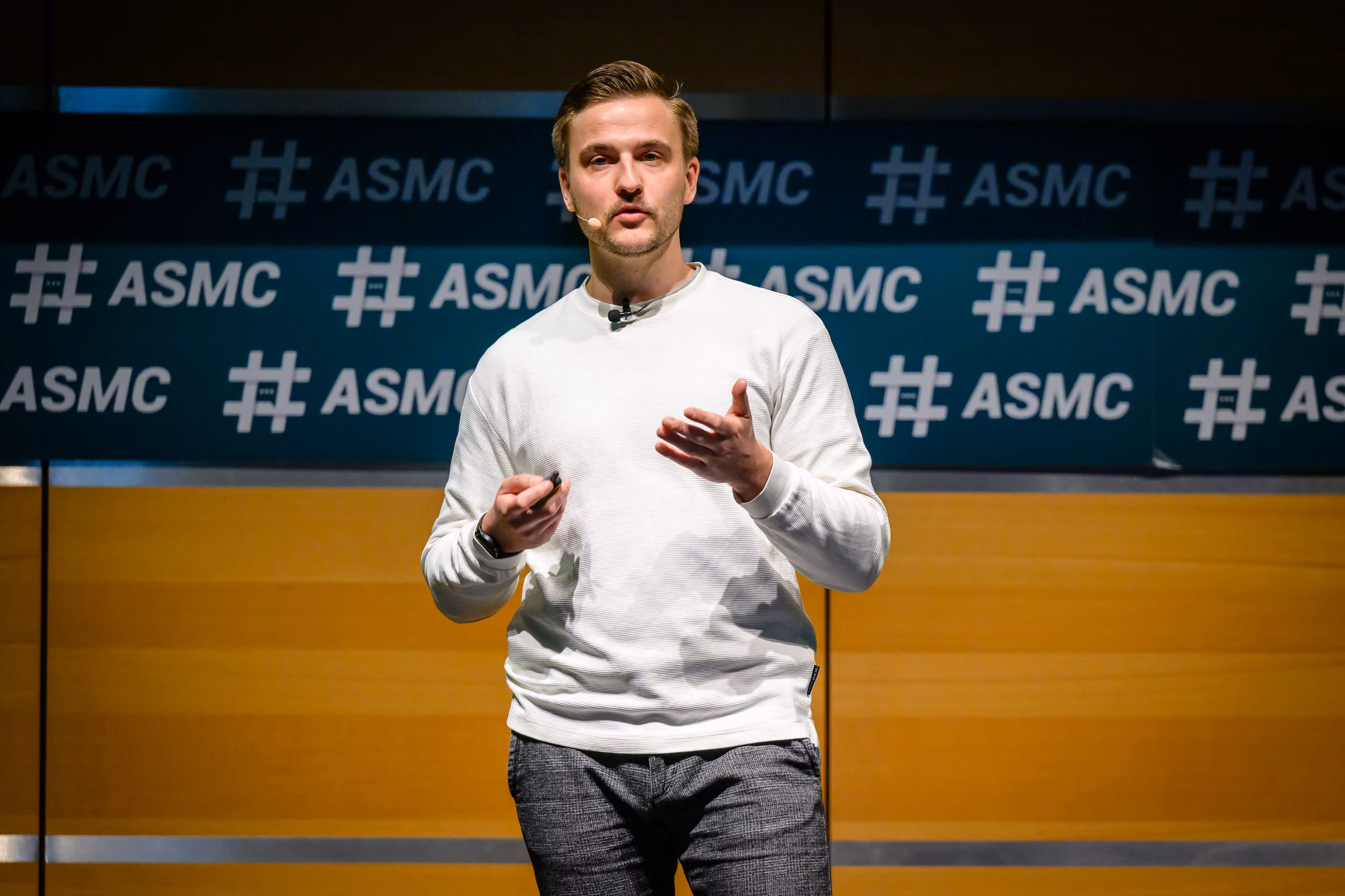 Ein Mann im weißen Pullover spricht mit Headset-Mikrofon vor einem ASMC-Logo-Hintergrund.