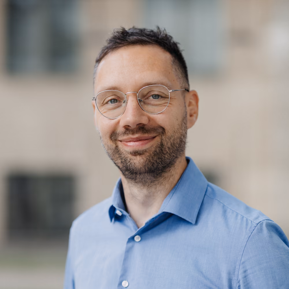 Porträt eines lächelnden Mannes mit Brille und blauem Hemd vor verschwommenem Hintergrund.