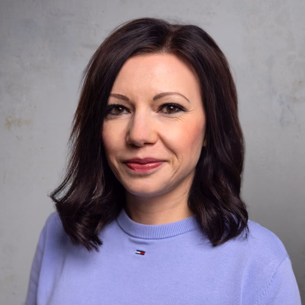 Frau mit mittellangen dunklen Haaren und einem hellvioletten Pullover vor grauem Hintergrund.
