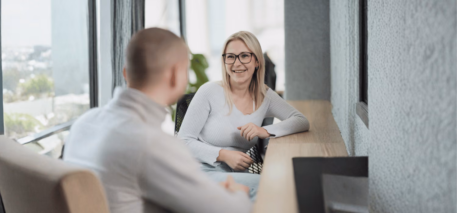 Zwei Menschen unterhalten sich an einem Holztisch in einem modernen hellen Büro mit Fensterblick.