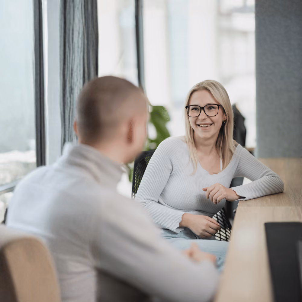 Zwei Personen unterhalten sich an einem hellen Bürofenster, eine lächelnde Frau mit Brille sitzt am Tisch.