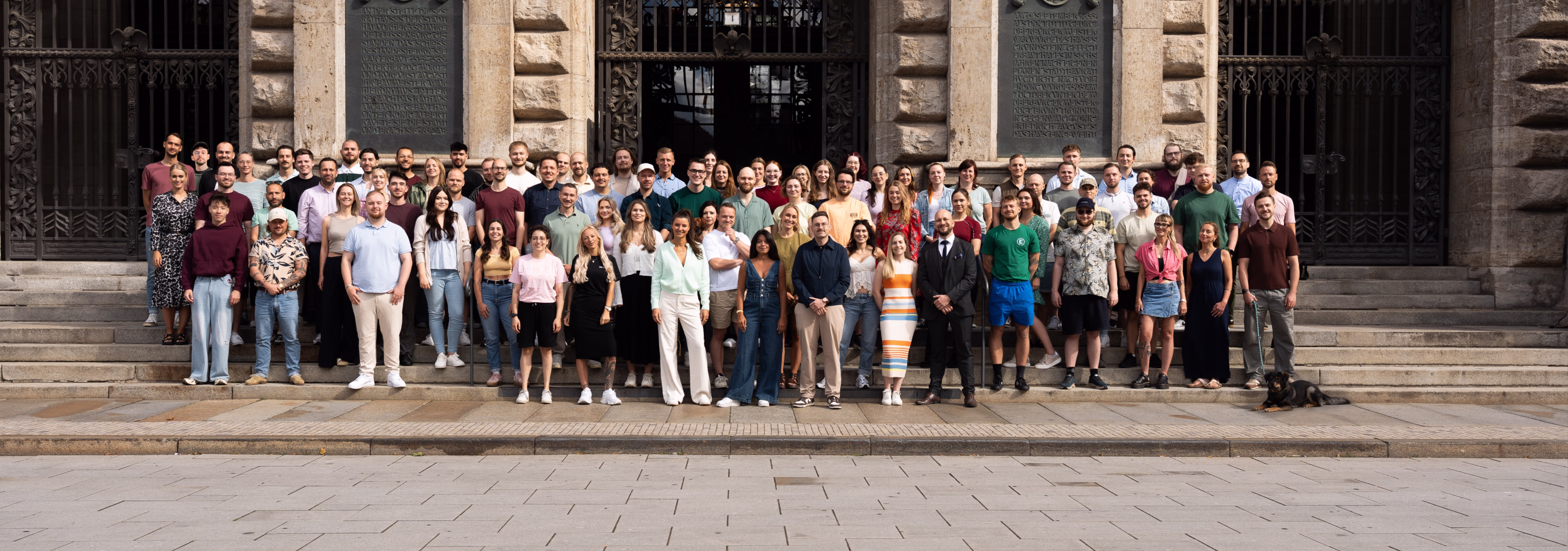 Großes Team von Männern und Frauen steht auf Stufen vor einem historischen Gebäude mit steinernen Säulen.