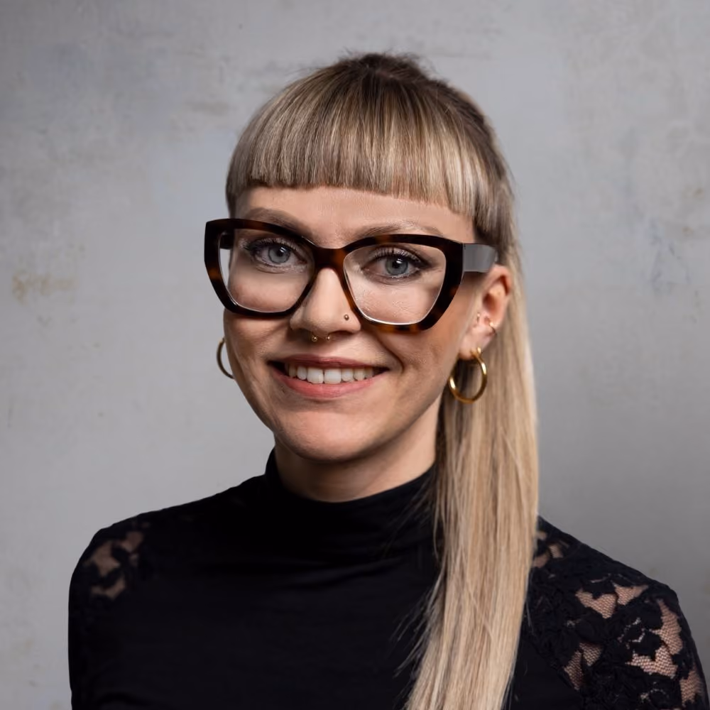 Porträt einer lächelnden Frau mit langen blonden Haaren, Pony, großen, dunklen Brillen und mehreren goldenen Ohrringen sowie Gesichtspiercings. Sie trägt ein schwarzes, langärmeliges Oberteil mit Spitzenmuster.