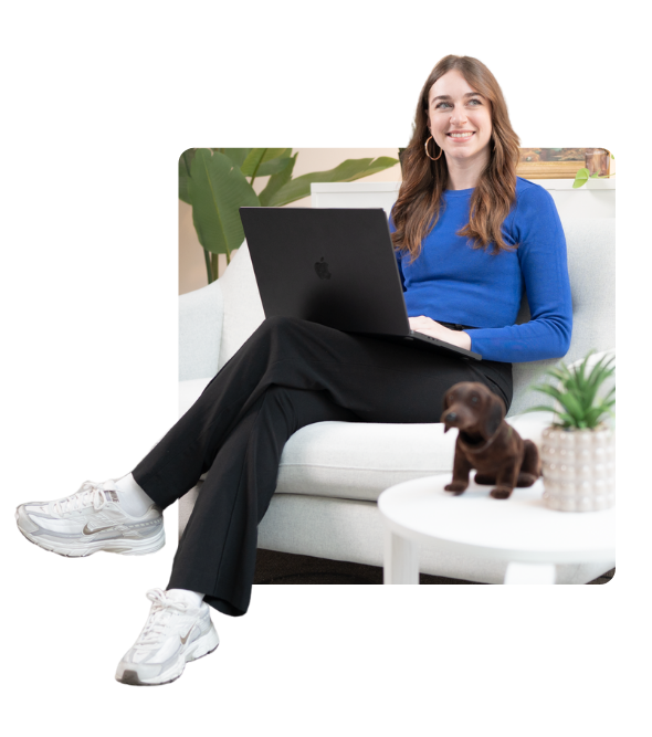 Eine lächelnde Frau mit langen braunen Haaren, die einen blauen Pullover, schwarze Hosen und weiße Turnschuhe trägt, sitzt auf einem weißen Sofa mit einem Laptop auf dem Schoß. Neben ihr steht ein runder weißer Tisch mit einer kleinen Pflanzen- und einer Dackel-Statue. Im Hintergrund sind grüne Pflanzblätter sichtbar.
